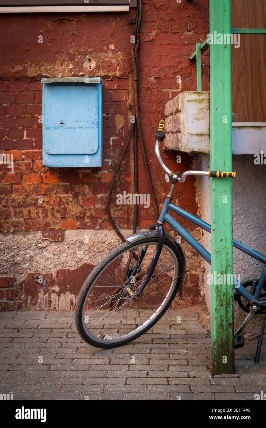 Blaues Fahrrad auf grünem Pfosten neben abblätternder roter Backsteinwand und pastellblauem Briefkasten, urbane Textur und Vintage-Straßenszene, rustikale Farben und abgenutzte Details. Stockfoto