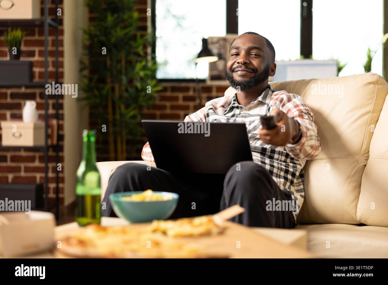 afroamerikanischer Freelancer arbeitet ferngesteuert vom Sofa aus mit einem digitalen Gerät auf dem Schoß und stellt die Lautstärke des Fernsehers über die Fernbedienung ein. Junger Mann Multitasking zwischen Laptop und Fernsehen zu Hause. Stockfoto