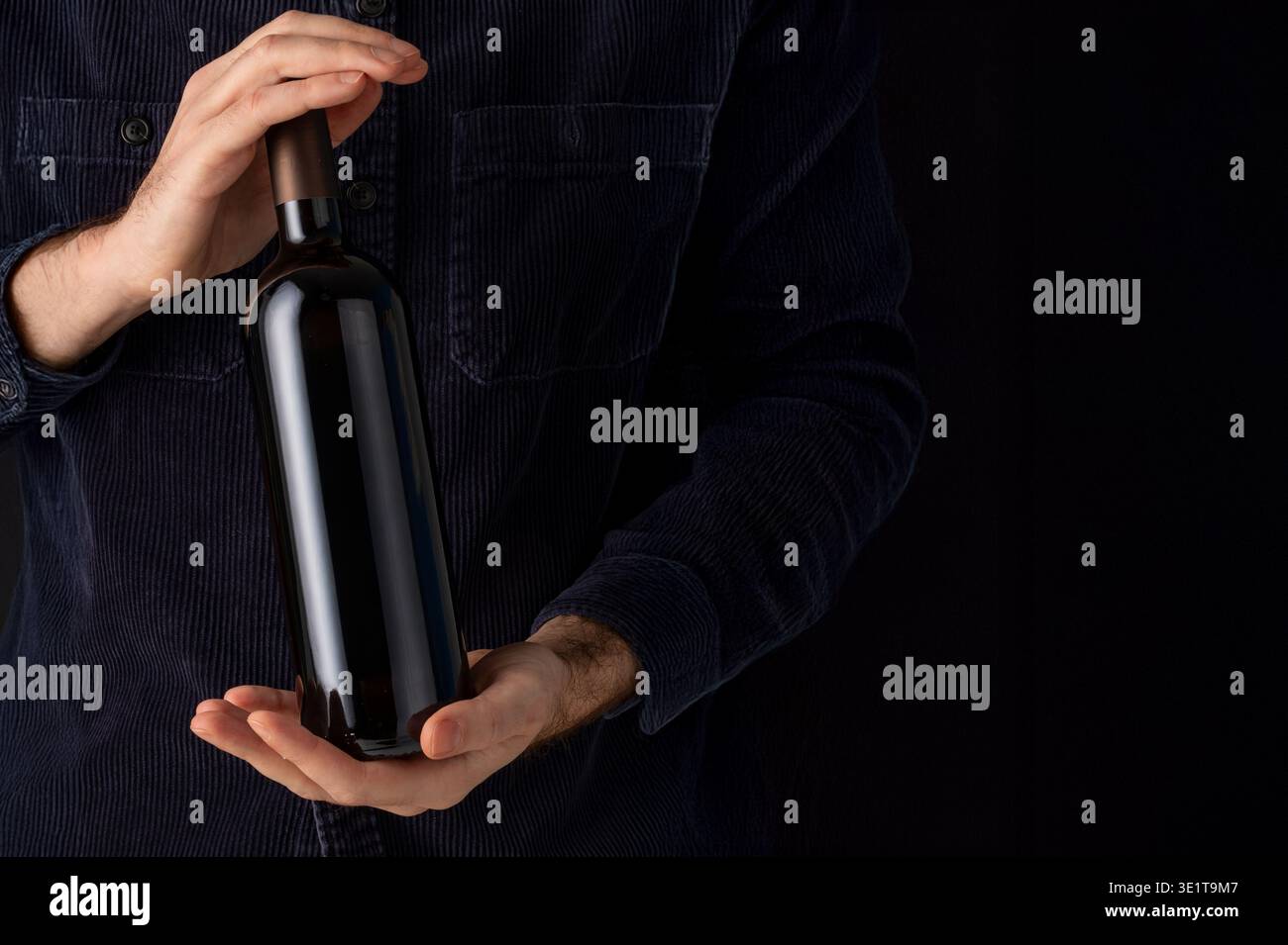 Mann blaues Samt Hemd hält Weinflasche schwarzes Etikett dunkler Hintergrund elegante Weingut Präsentation Keller Stockfoto