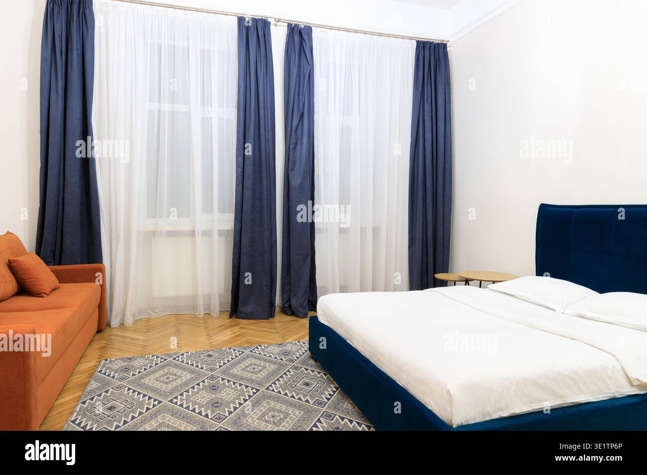 Ein stilvolles und komfortables Schlafzimmer mit einem weichen orangen Sofa, einem tiefblauen Polsterbett und einem geometrisch gemusterten Teppich auf Holzboden Stockfoto
