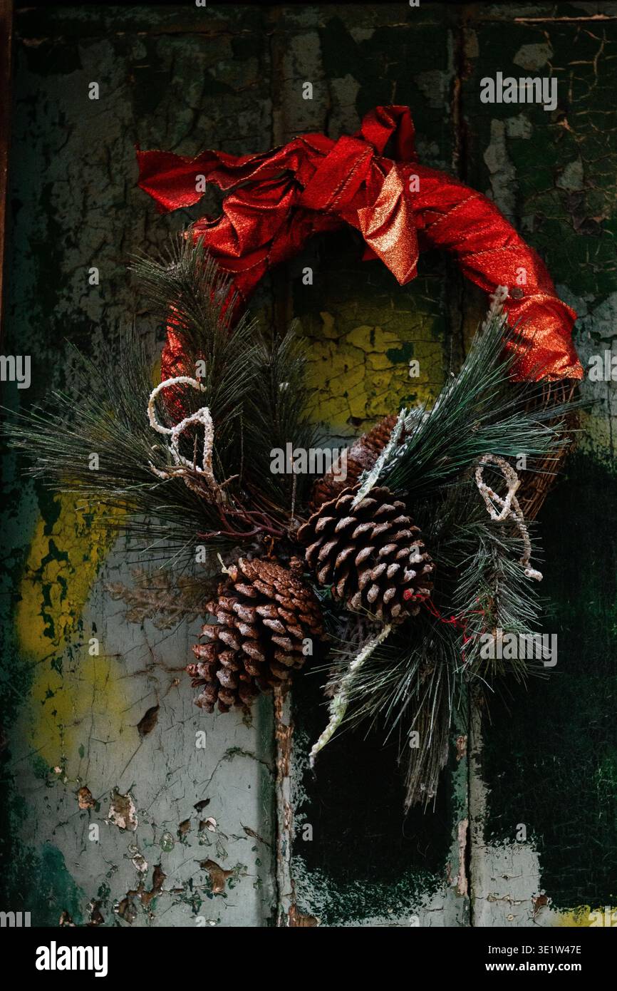Ein rotes Band ziert einen Weihnachtskranz aus Tannenzapfen und Nadeln an einer alten, abblätternden, bemalten Tür. Stockfoto
