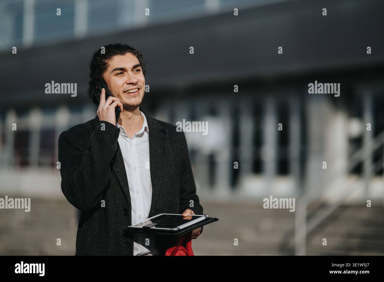 Multikultureller Geschäftsmann, der am Telefon telefoniert, während er ein Tablet vor einem modernen Bürogebäude hält Stockfoto