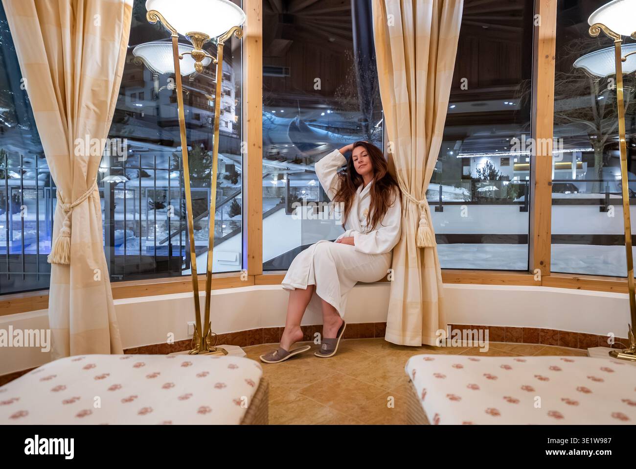 Frau im Spa-Bademantel am gebogenen Erkerfenster im alpinen Hotel nachts Stockfoto
