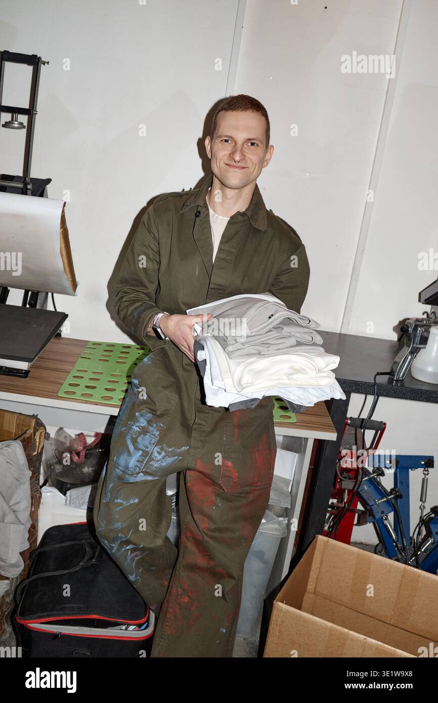 Vertikal des jungen Erwachsenen, der gefaltete Hemden im industriellen Arbeitsbereich hält Stockfoto