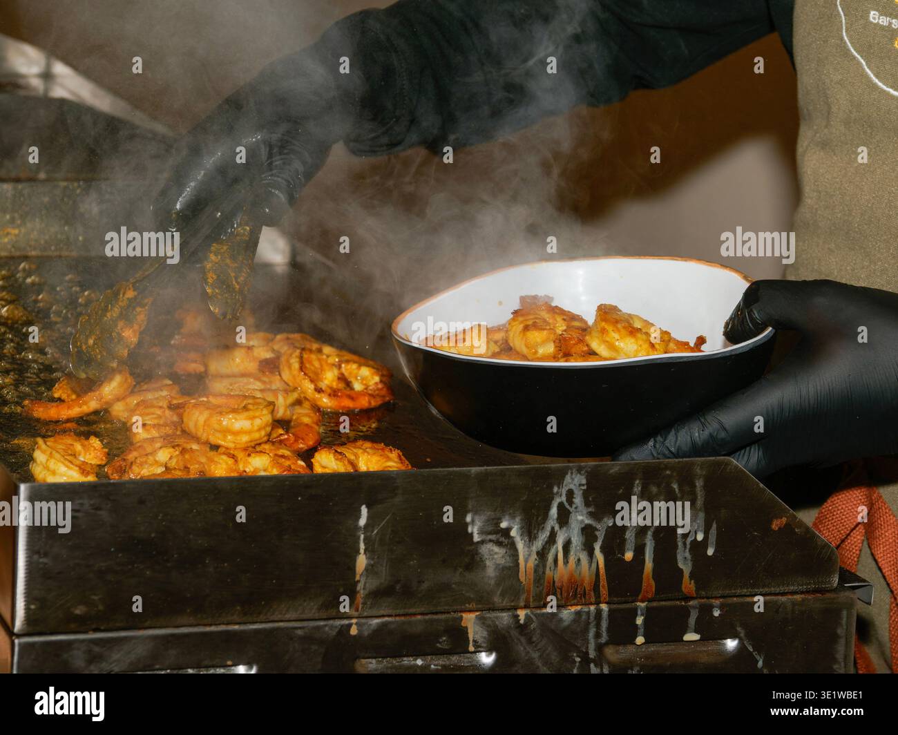 Nahaufnahme gegrillter Garnelen, die auf einem heißen Grill gekocht werden, wobei ein Koch schwarze Handschuhe trägt und frisch gegrillte Meeresfrüchte in einer Schüssel serviert. Stockfoto