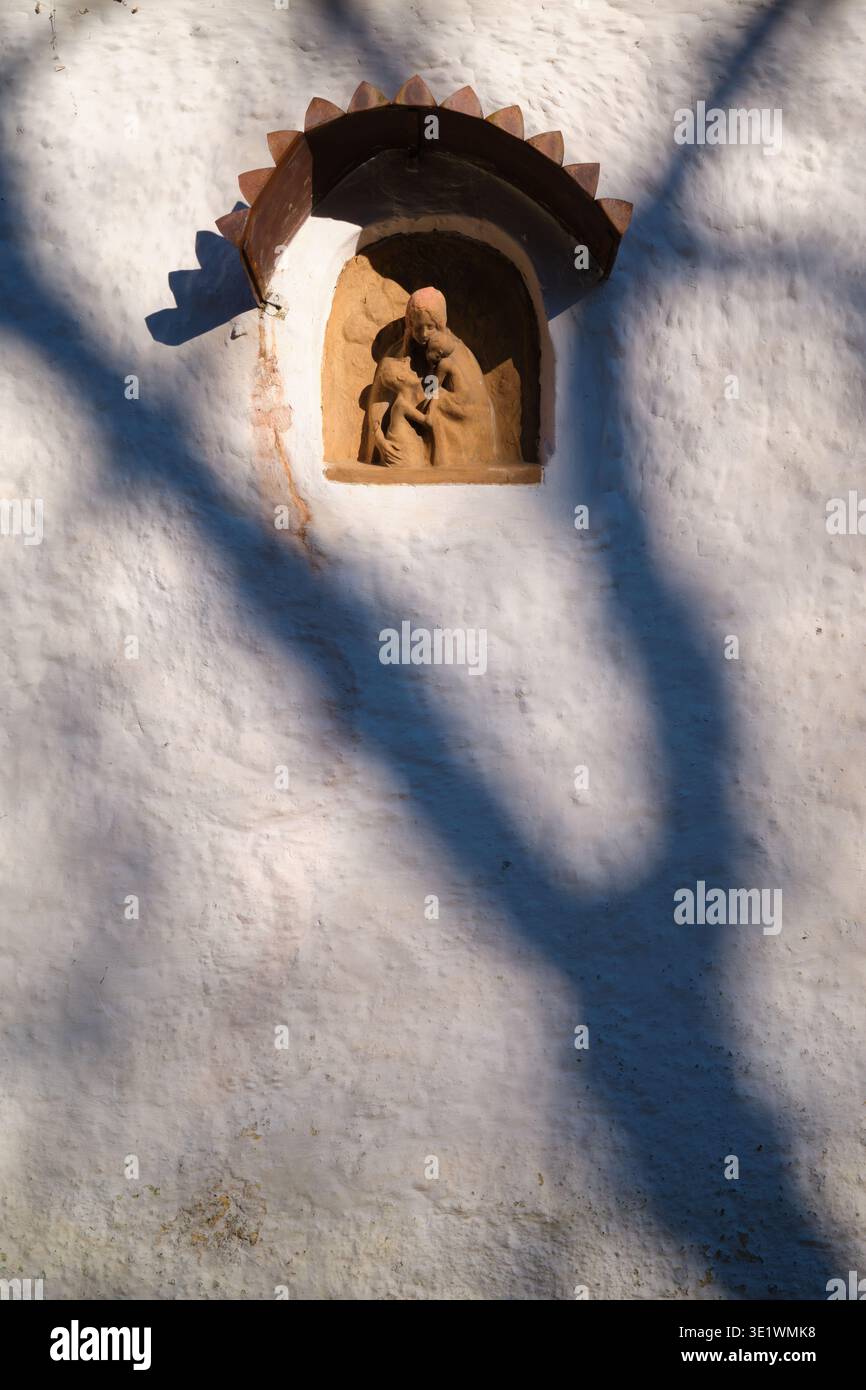 Kleine religiöse Skulptur einer Mutter und eines Kindes in einer Wandnische, beleuchtet von warmem Sonnenlicht mit dramatischen Baumschatten über einer strukturierten Wand Stockfoto