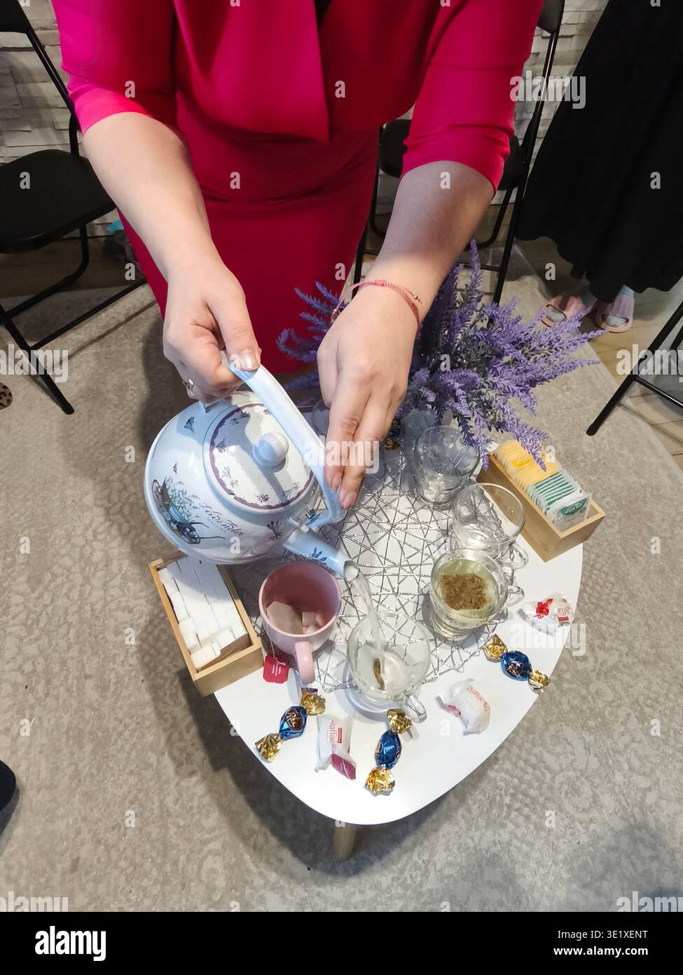 Frau gießt heißen Tee an einem gemütlichen Tisch mit Tassen, Süßigkeiten und Lavendeldekoration, entspannender geselliger Moment drinnen. Stockfoto