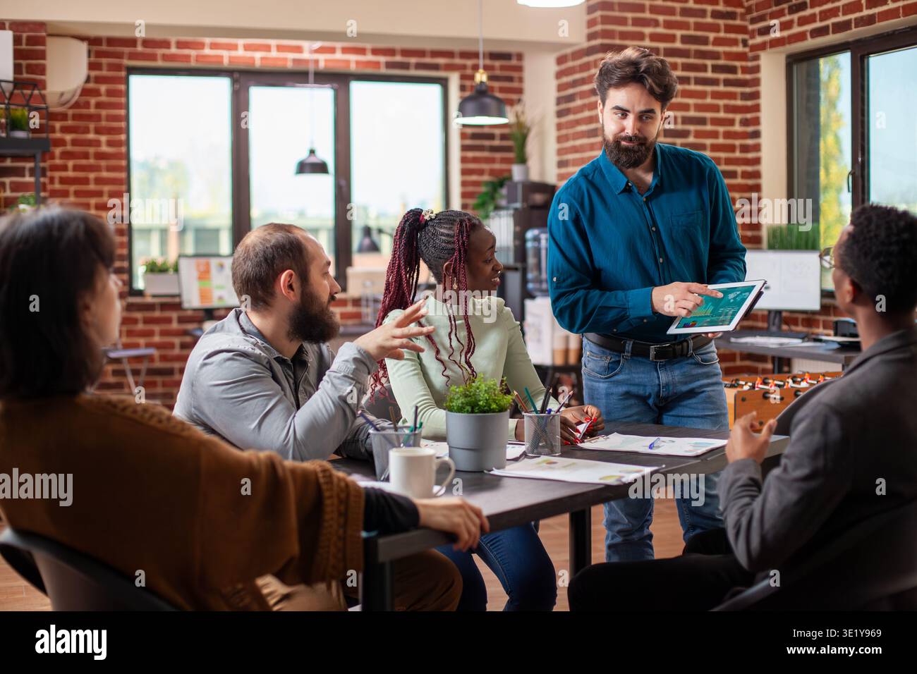 Weißbärtiger Mann, der mit einem Tablet steht und Projektaktualisierungen mit Kollegen in modernen Büros teilt. Das vielfältige Startup-Team hört den männlichen Analysten aufmerksam zu, die bei Gruppenbesprechungen finanzielle Präsentationen machen Stockfoto