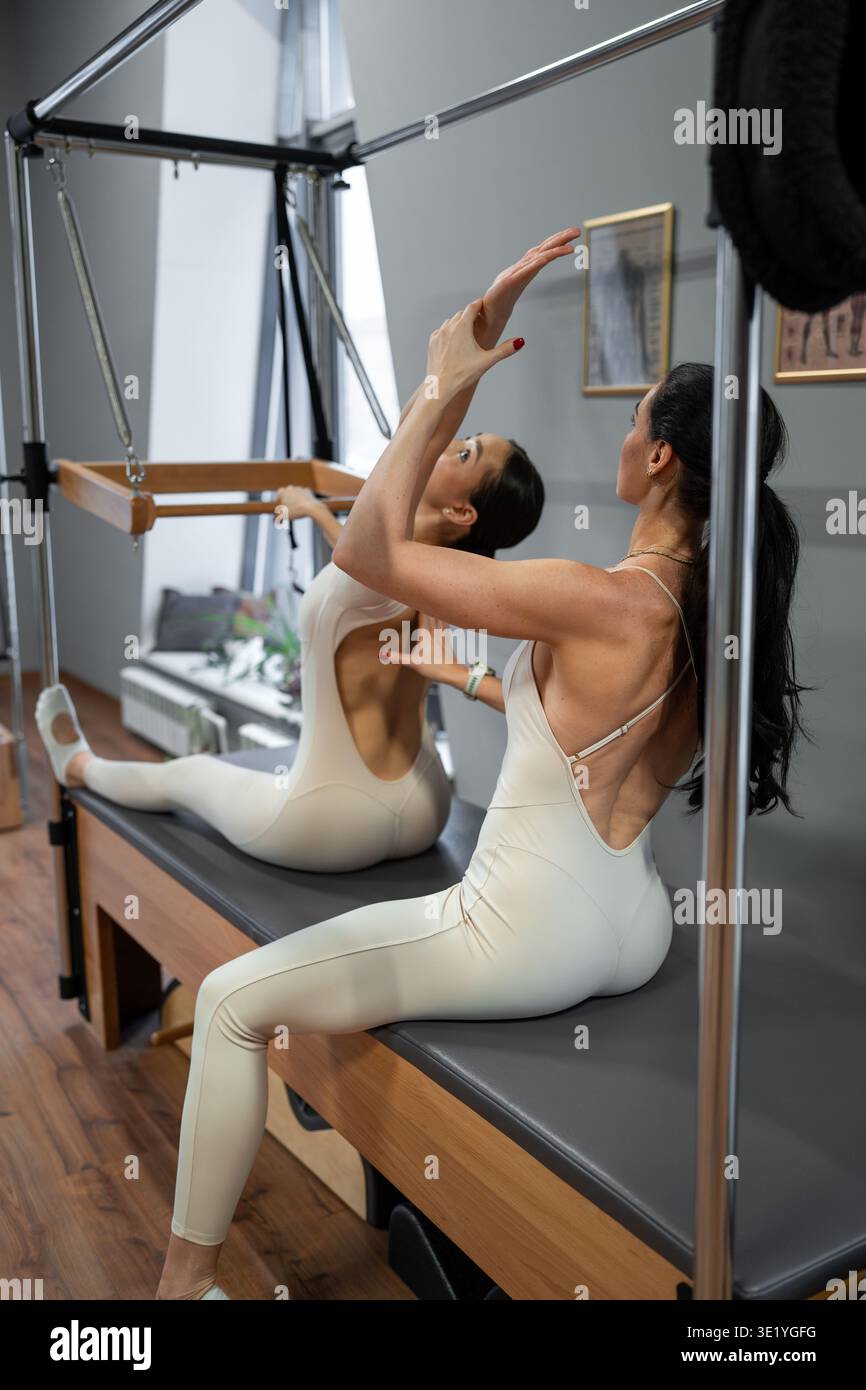 Pilates-Duo unterstützt sich gegenseitig mit Flexibilität und Balance auf den Geräten in hellen Fitnessräumen. Assistieren, Flexibilität, Pilates, Balance, Duo Stockfoto