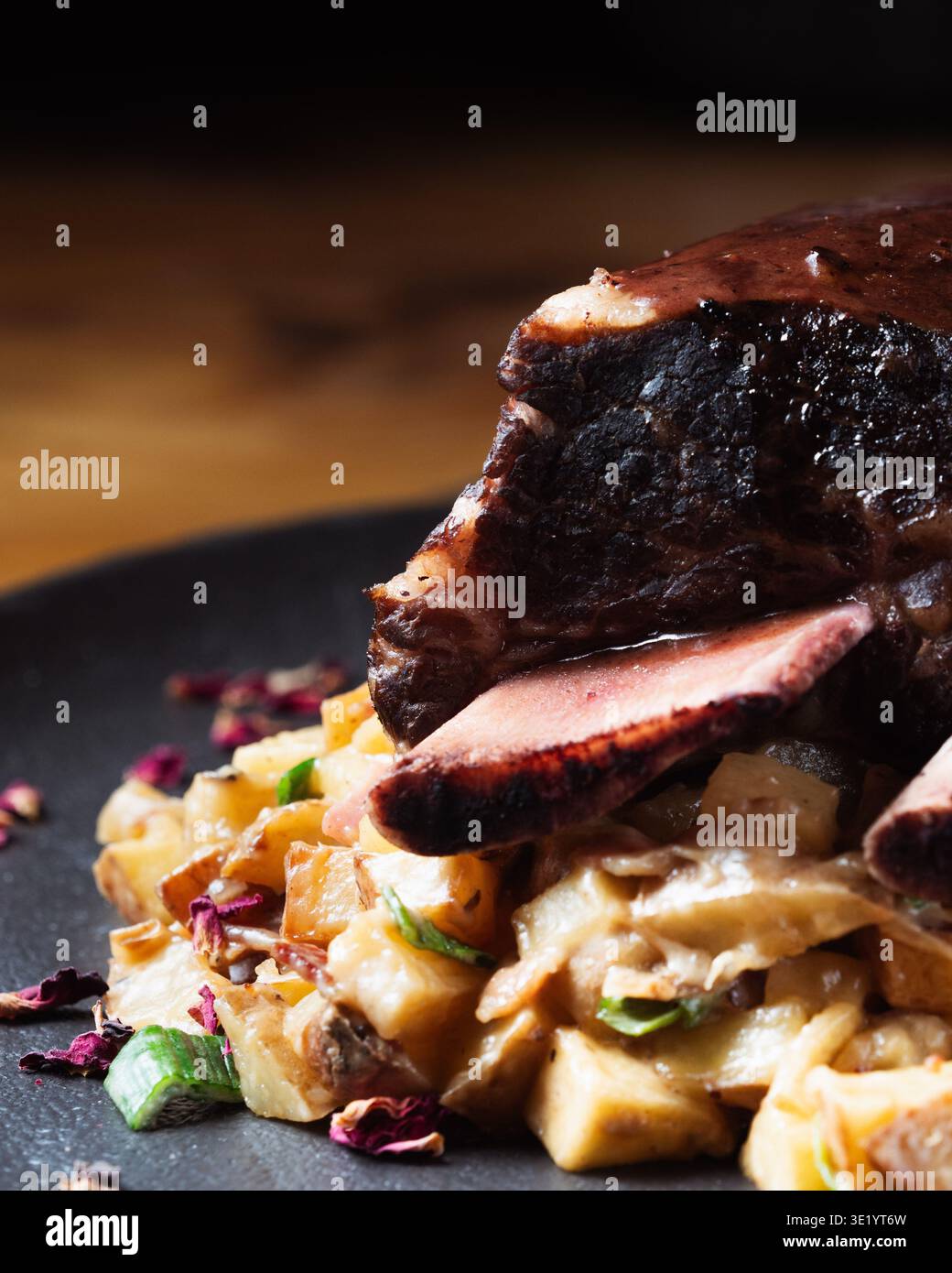 Reichhaltiges, dunkles Fleisch liegt auf cremigem Kartoffelbrei. Getrocknete Rosenblätter verleihen Farbe und Eleganz. Garniert mit gehackten grünen Kräutern sieht das Gericht dekadent aus. Stockfoto