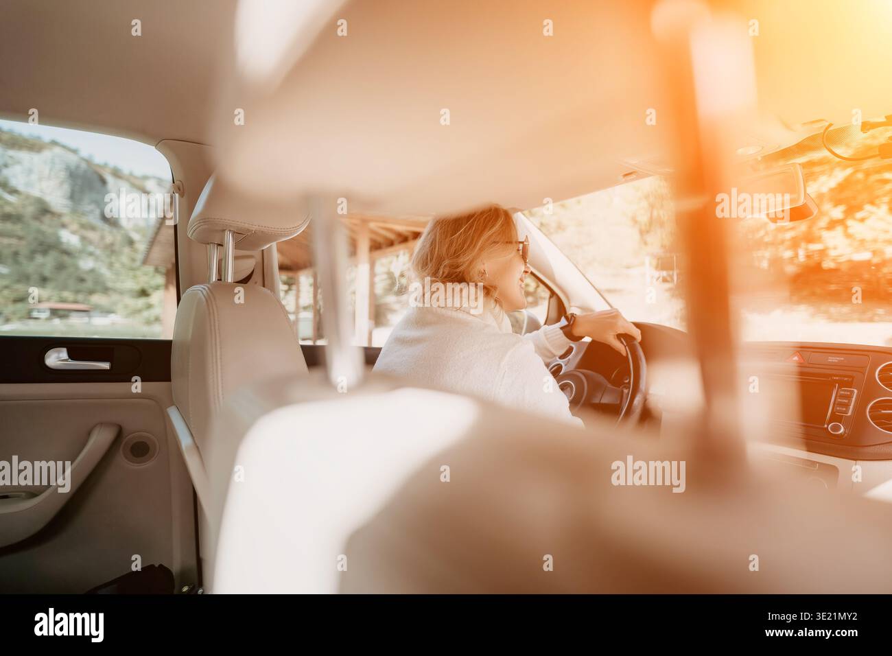 Frau, die Auto-Rücksitz-Sonne fährt – Eine Frau fährt ein Auto, während sie auf dem Rücksitz sitzt, während die Sonne durch das Fenster scheint. Stockfoto