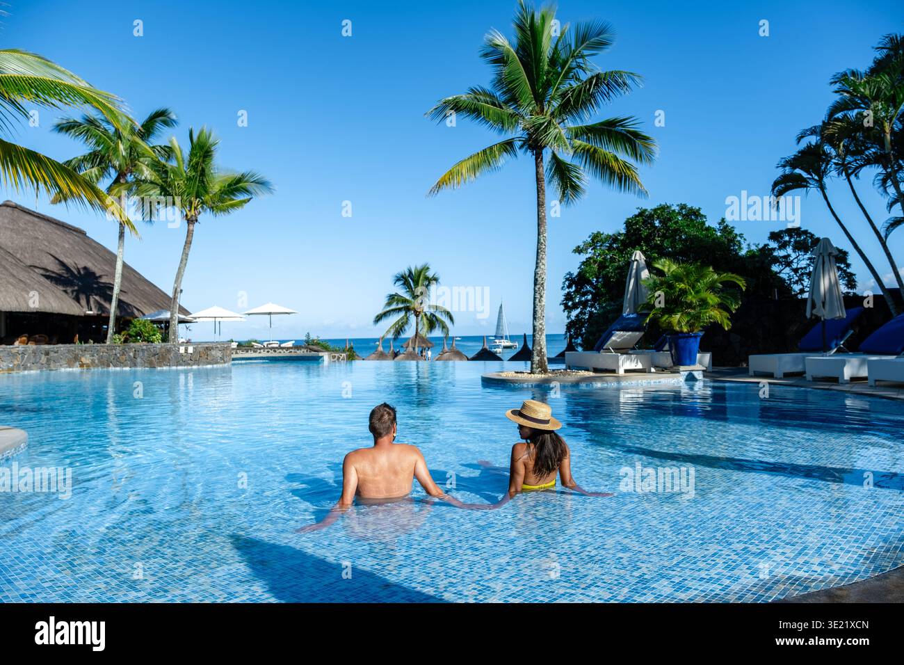 Paare entspannen sich in einem atemberaubenden Infinity-Pool umgeben von Palmen und genießen die tropische Brise und den atemberaubenden Meerblick auf Mauritius. Ein perfekter Rückzugsort für Romantik und Freizeit. Stockfoto