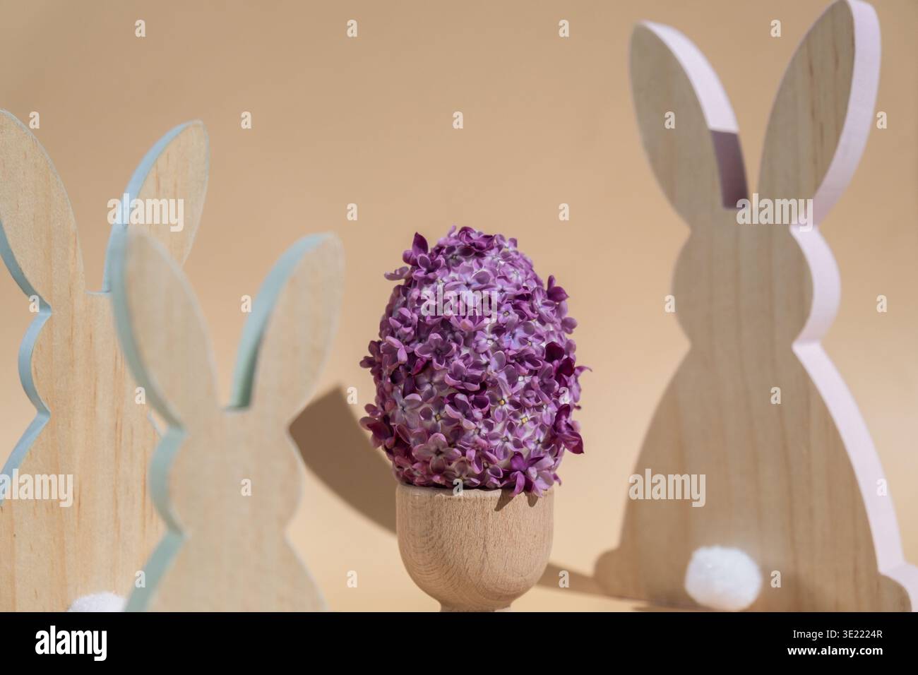 Minimalistisches, dezentes Hasendesign aus Holz. Osterei aus Flieder-Frühlingsblumen in pastellbeigem Hintergrund. Nachhaltiger Urlaub minimalistische Stillleben-Komposition. Raumvorlage für Einladungsgrußkarte Postkarte kopieren Stockfoto