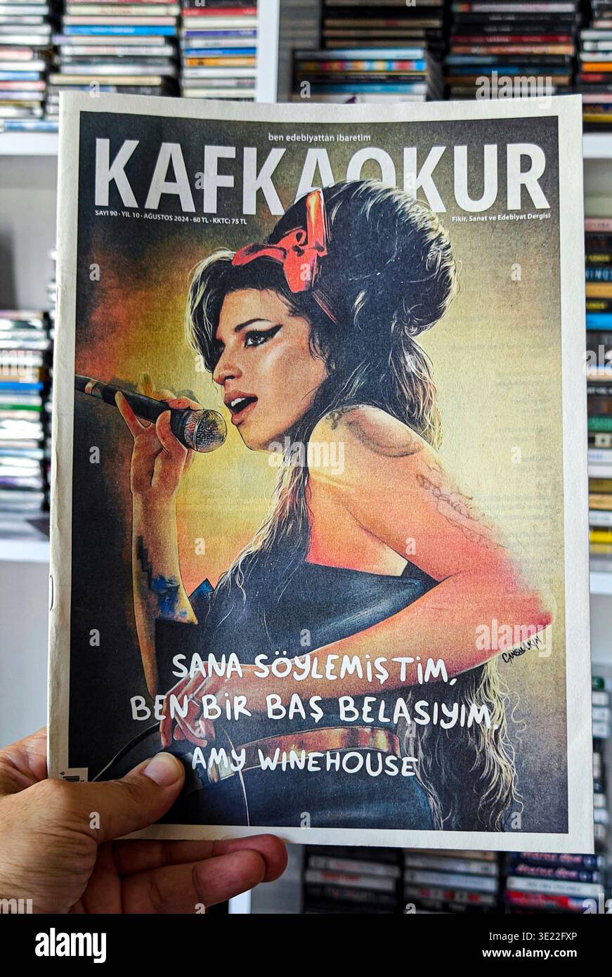 Hand hält eine Kopie des türkischen Literaturmagazins KAFKAOKUR mit einem Porträt von Amy Winehouse gegen eine verschwommene CD-Sammlung Stockfoto