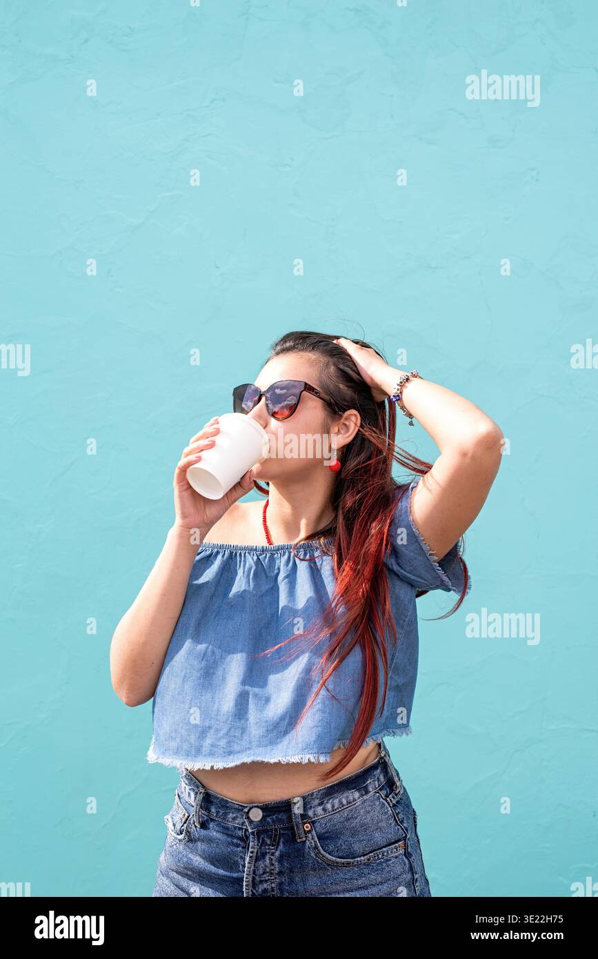 Attraktive junge Frau in Sommerkleidung und Sonnenbrille, die eine Tasse Kaffee in den Händen hält, auf blauem Hintergrund auf der Straße tanzt || Model veröffentlicht Stockfoto