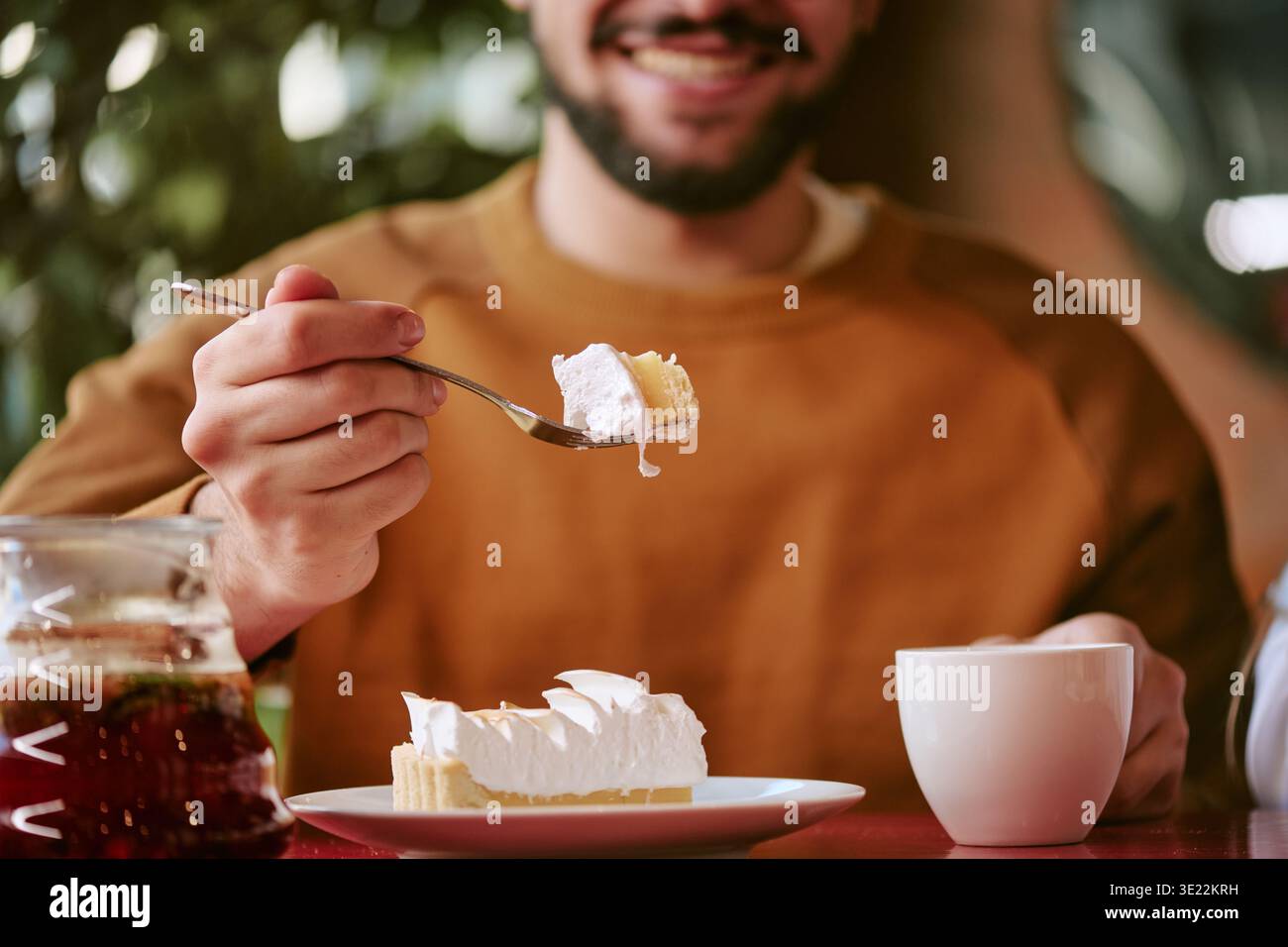 Junger erwachsener Mann lächelt, während er Kuchen am Cafe Table isst Stockfoto