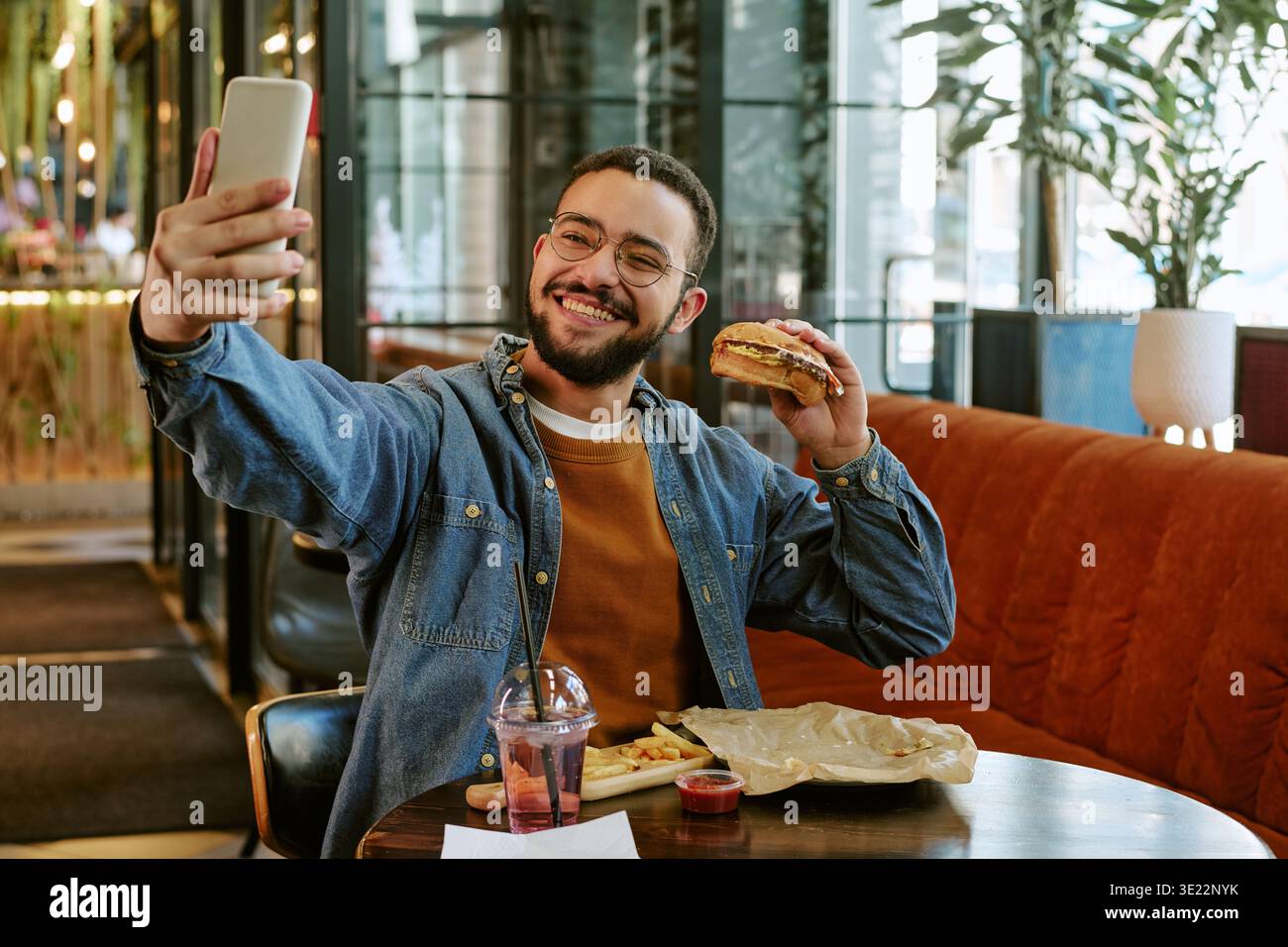 Junger Erwachsener, der Selfie nimmt, während er Burger im Restaurant isst Stockfoto