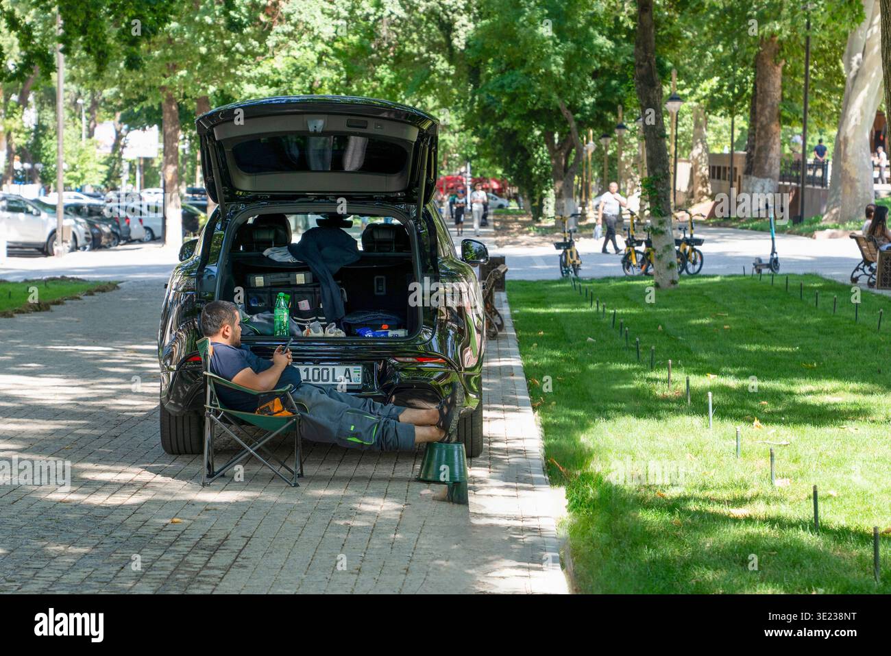 Taschkent (Toschkent), Usbekistan (O'zbekiston) - 26. Mai 2025: Ein einheimischer Mann, der sich in einem Campingstuhl neben seinem Auto in einem üppig grünen Stadtpark entspannt und einen genießt Stockfoto
