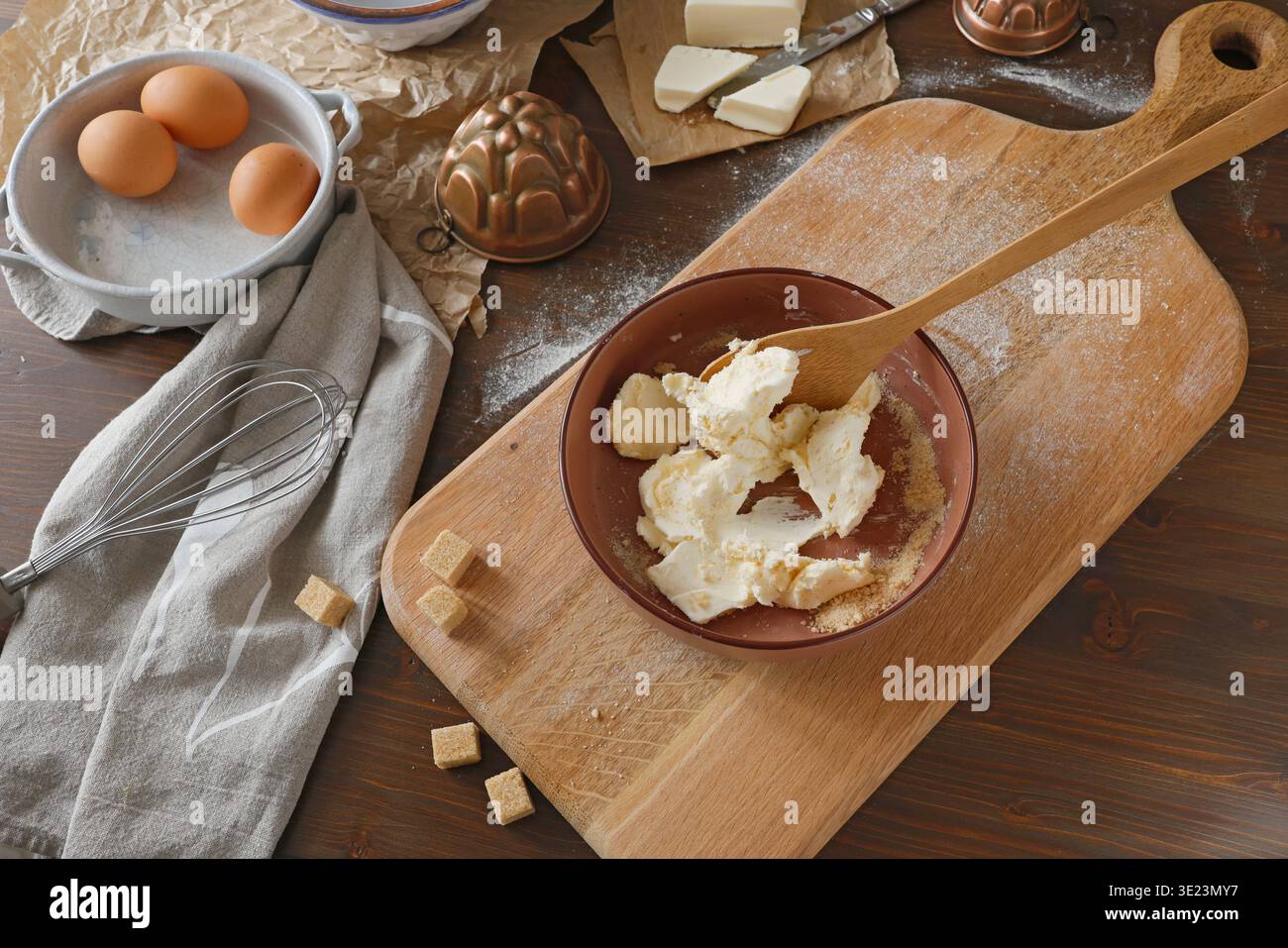 Butter und Zucker in einer Schüssel zum Backen auf rustikalem Holztisch mischen. Hochwinkelaufnahme von Kuchenzutaten einschließlich frischer Eier, Schneebesen, Butter und Kupfer Stockfoto