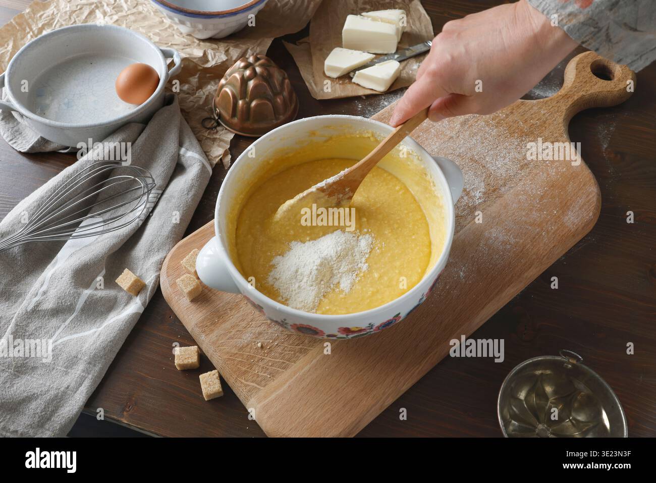 Mehl in Kuchenteig in einer Keramikschale auf Holzbrett mischen. Nahaufnahme der Hand mit einem Holzlöffel, der Teig mit Zutaten wie Eiern und zubereitet Stockfoto