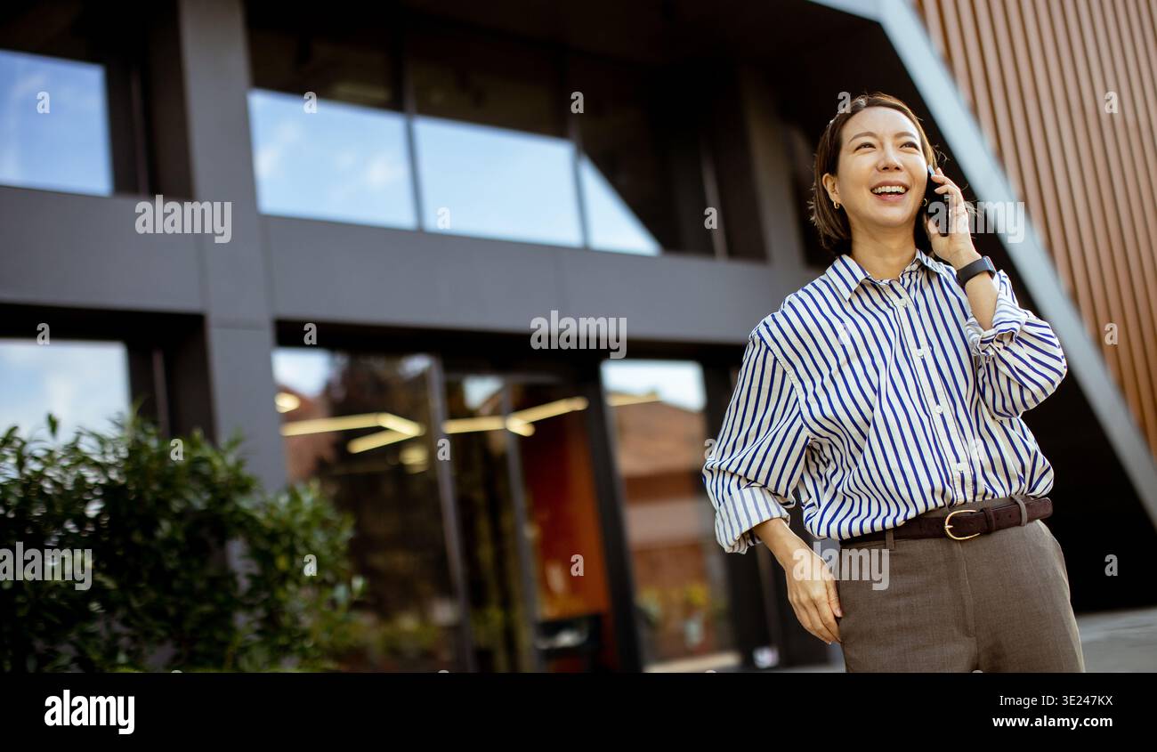 Eine Koreanerin steht vor einem modernen Gebäude. Sie lächelt, während sie am Telefon spricht. Der Himmel ist klar, und hinter ihr sind große Fenster. Stockfoto