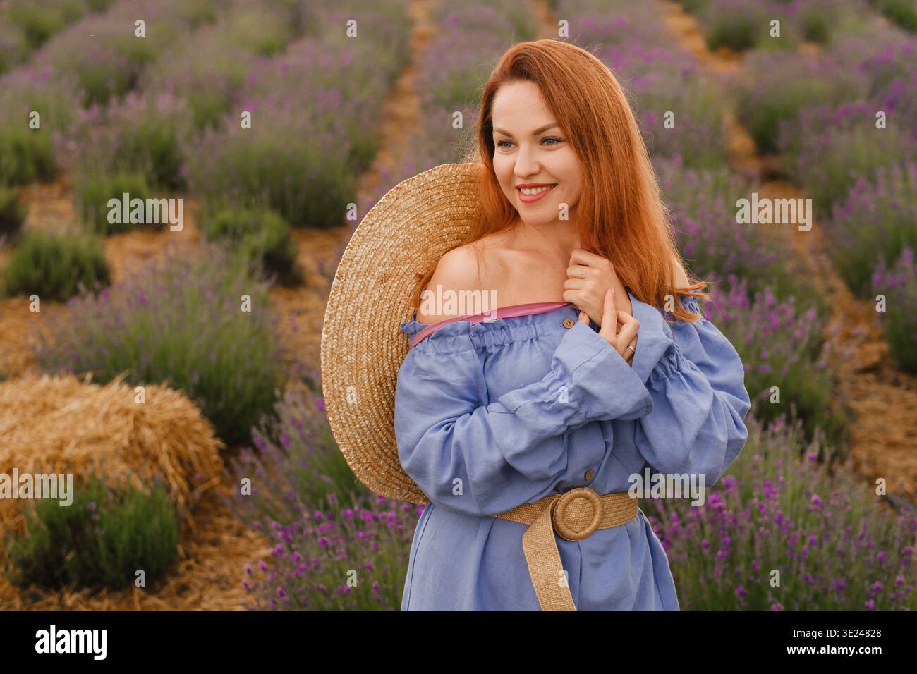 Eine Frau steht in einem Lavendelfeld, trägt ein blaues Kleid mit einem breitkrempigen Strohhut. Sie lächelt und genießt das sonnige Wetter in einer ländlichen Gegend Stockfoto