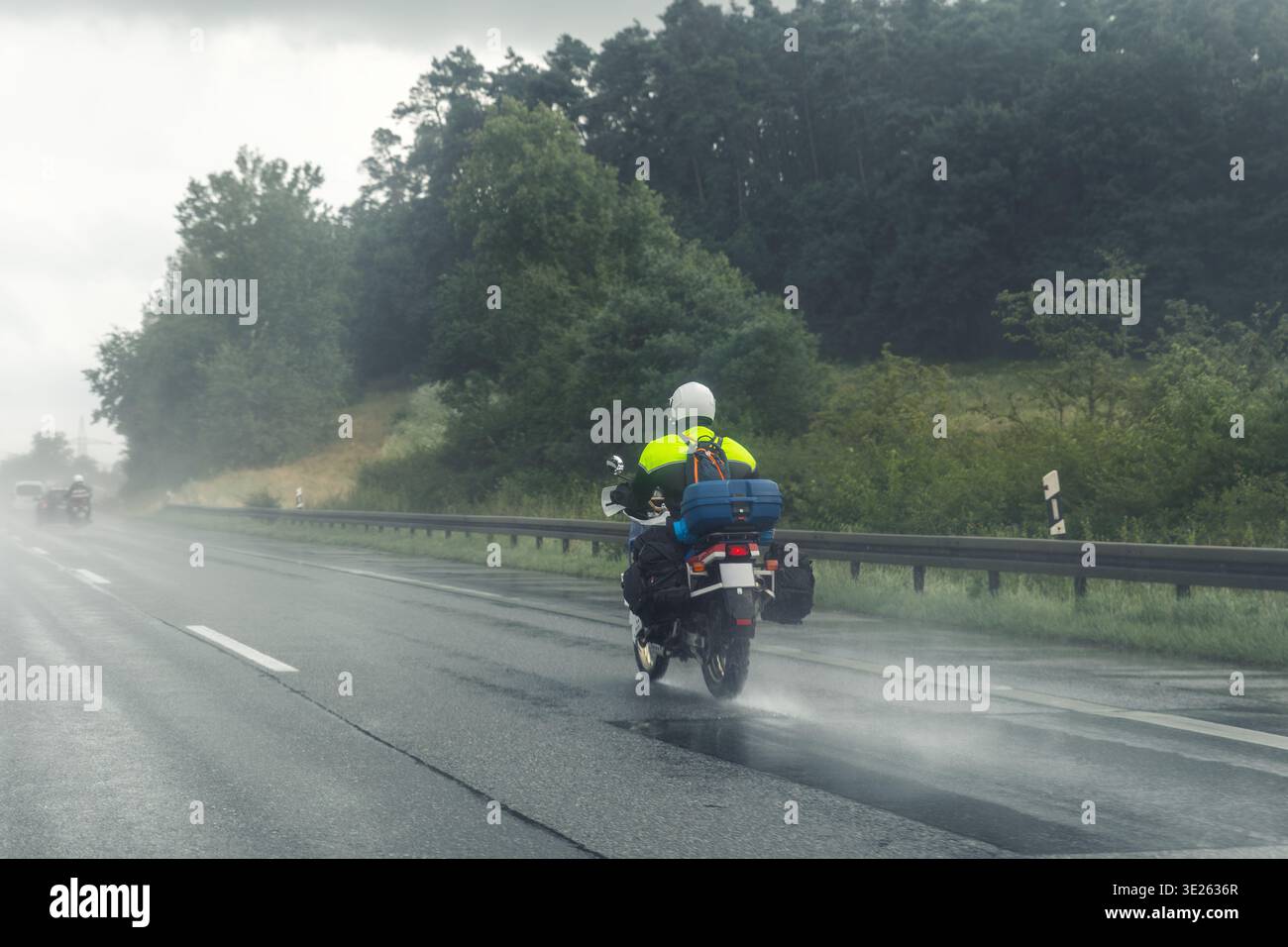 Motorradfahrer fahren auf nasser Autobahn im Regenschauer mit geringer Sicht umgeben von grünem Waldnebel. Die reflektierende Jacke für Tourenreiter reist stürmisch Stockfoto