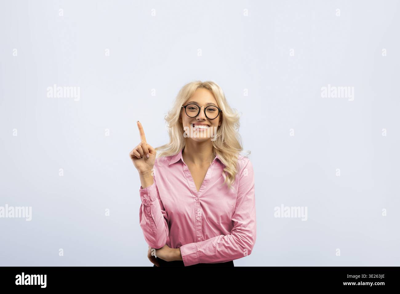 Frau lächelt und zeigt nach oben, während sie drinnen vor einem Ebenen Hintergrund steht Stockfoto