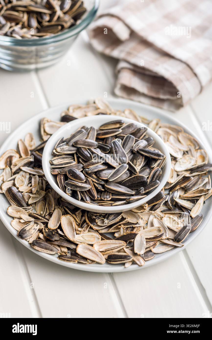 Eine Nahaufnahme einer weißen Schüssel gefüllt mit gerösteten Sonnenblumenkernen, umgeben von abgelegten Muscheln auf einem Teller, was auf lässiges Snacken, Textur und Rost hindeutet Stockfoto