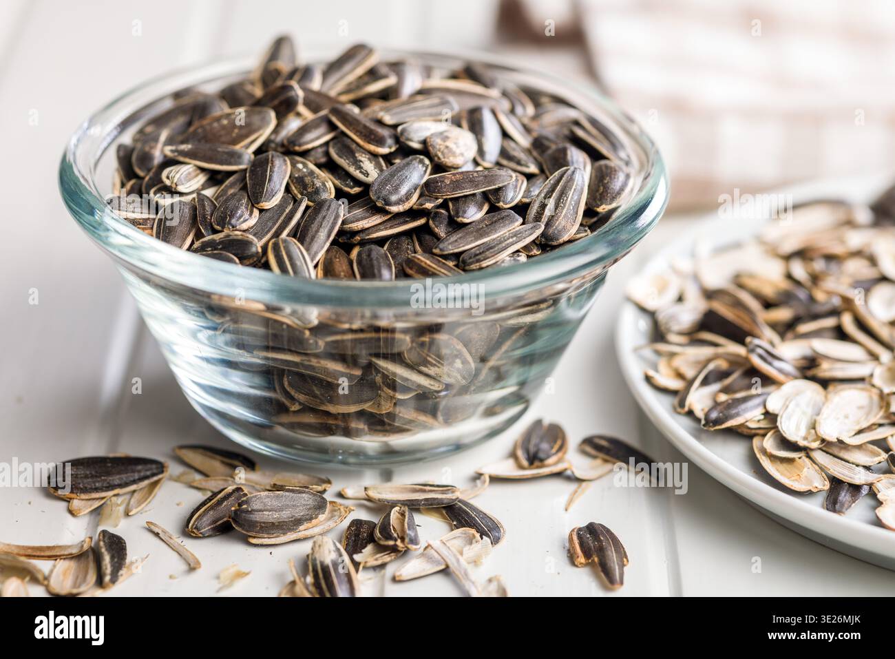Glasschale gefüllt mit Sonnenblumenkernen, umgeben von leeren Muscheln auf einem weißen Tisch, Nahaufnahme mit gestreiften Muscheln und knuspriger Textur A nat Stockfoto