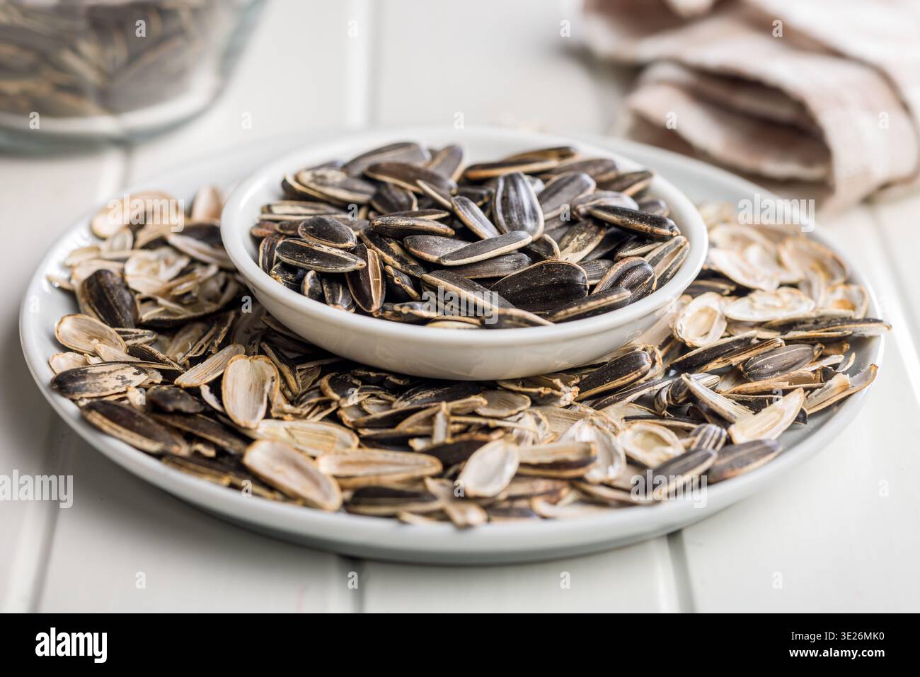 Eine Nahaufnahme einer weißen Schüssel gefüllt mit gerösteten Sonnenblumenkernen, umgeben von abgelegten Muscheln auf einem Teller, was auf lässiges Snacken, Textur und Rost hindeutet Stockfoto