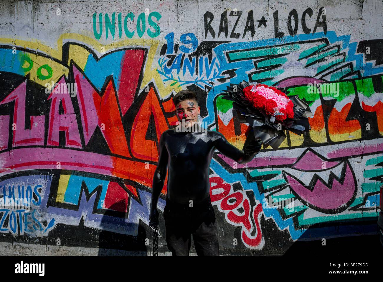 Mann malte schwarz mit roten Blumen, die während eines mexikanischen Karnevals vor einem farbenfrohen Graffiti-Wandgemälde posierten Stockfoto