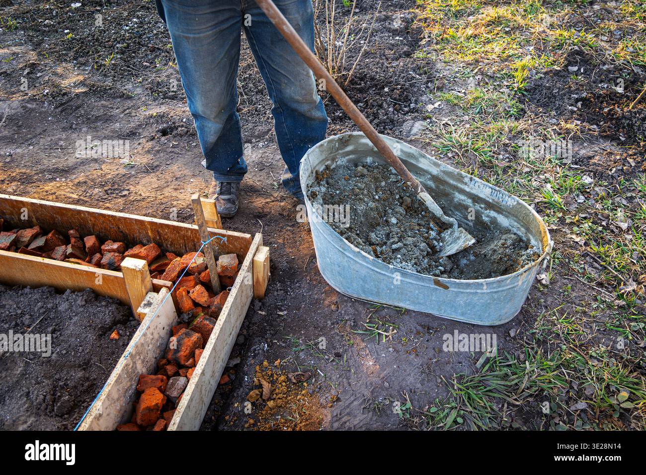 Mischbeton neben gemauertem Gartenfundament. Der Arbeiter schaufelt nassen Beton aus einer Metallwanne neben einem mit Ziegeln gefüllten Graben, um einen kleinen Außenbereich vorzubereiten Stockfoto