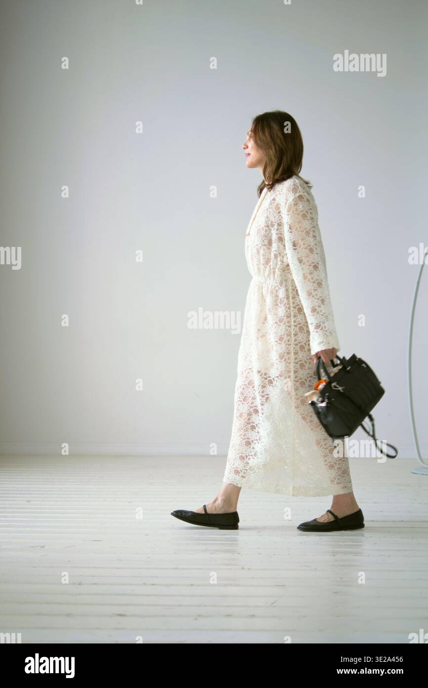 Frau in einem weißen Spitzenkleid hält eine schwarze Handtasche, während sie in einem hellen steht Stockfoto