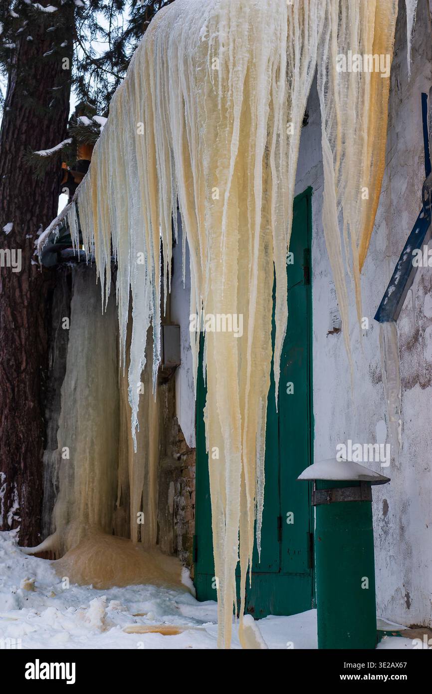 Riesige gelbliche Eiszapfen hängen dramatisch von einem verwitterten Garagendach ab und schaffen ein atemberaubendes, gefrorenes Spektakel. Die rohe Kraft des Winters verwandelt das Alltägliche Stockfoto