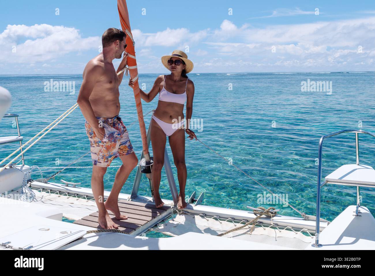 Ein stilvolles Paar entspannt sich auf einem Katamaran auf Mauritius, umgeben von atemberaubendem türkisfarbenem Wasser. Die helle Sonne erleuchtet ihre fröhlichen Ausdrucksformen, während sie den Luxus dieses tropischen Kurzurlaubs genießen. Stockfoto