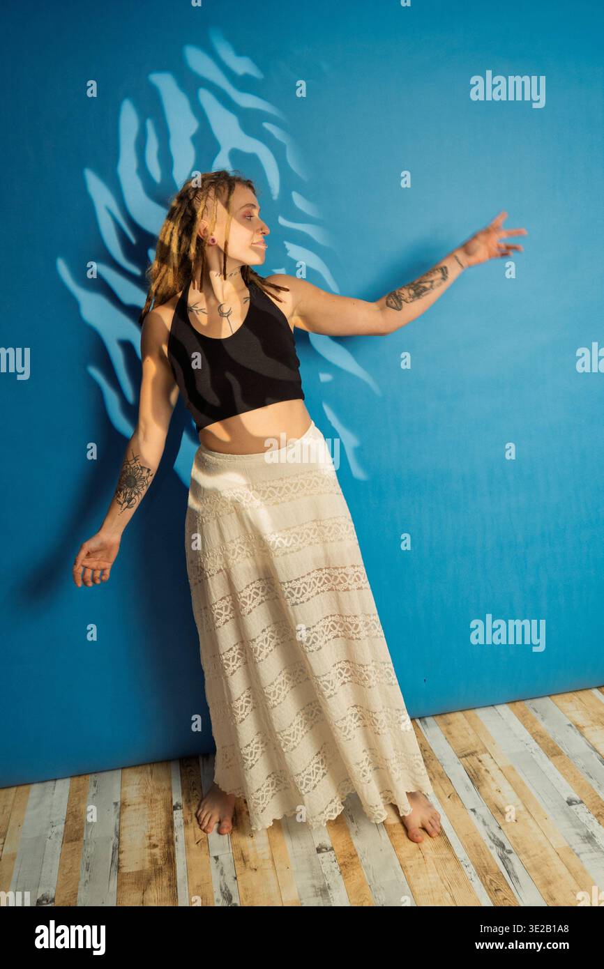 Junge Frau mit Tattoos und Dreadlocks, die in einem langen Rock vor blauem Hintergrund posiert Stockfoto