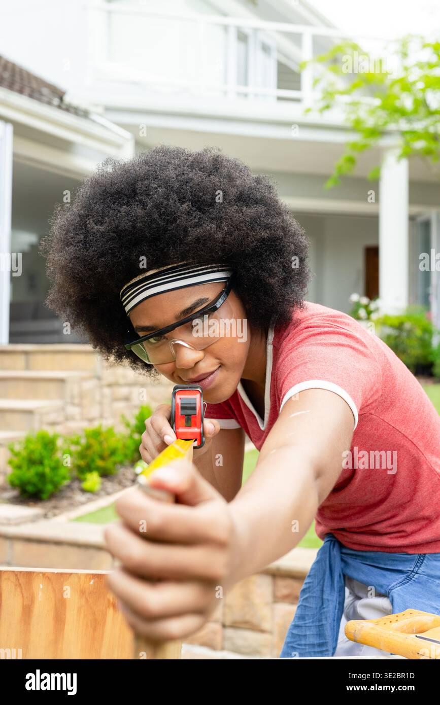 Afrikanische Frau mit Schutzbrille, die Holzdiele im Vorhof mit gelbem Klebeband misst. Holzbearbeitung, diy, Outdoor, Vorort, Werkzeuge, Klemme, Veranda Stockfoto