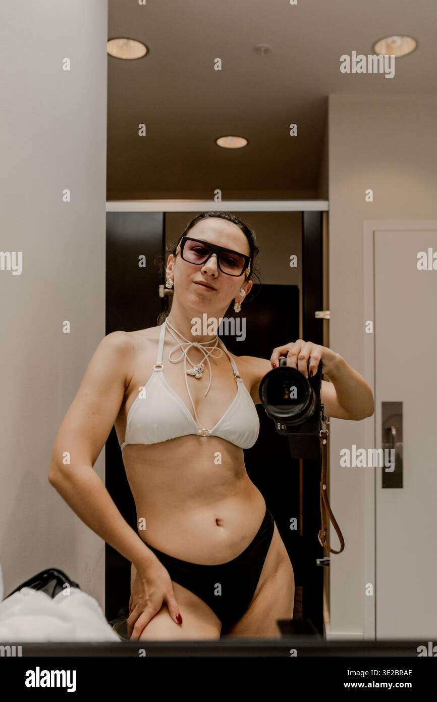 Frau steht vor dem Spiegel und macht ein Foto, während sie in einem Badezimmer mit neutralen Farben und sanfter Beleuchtung einen Bikini und eine Brille trägt Stockfoto