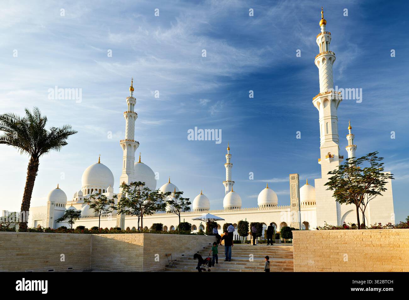 Die Menschen laufen in der Nähe der wunderschönen Architektur der Scheich-Zayed-Moschee in Abu Dhabi. Der Himmel ist tagsüber klar und blau. Stockfoto