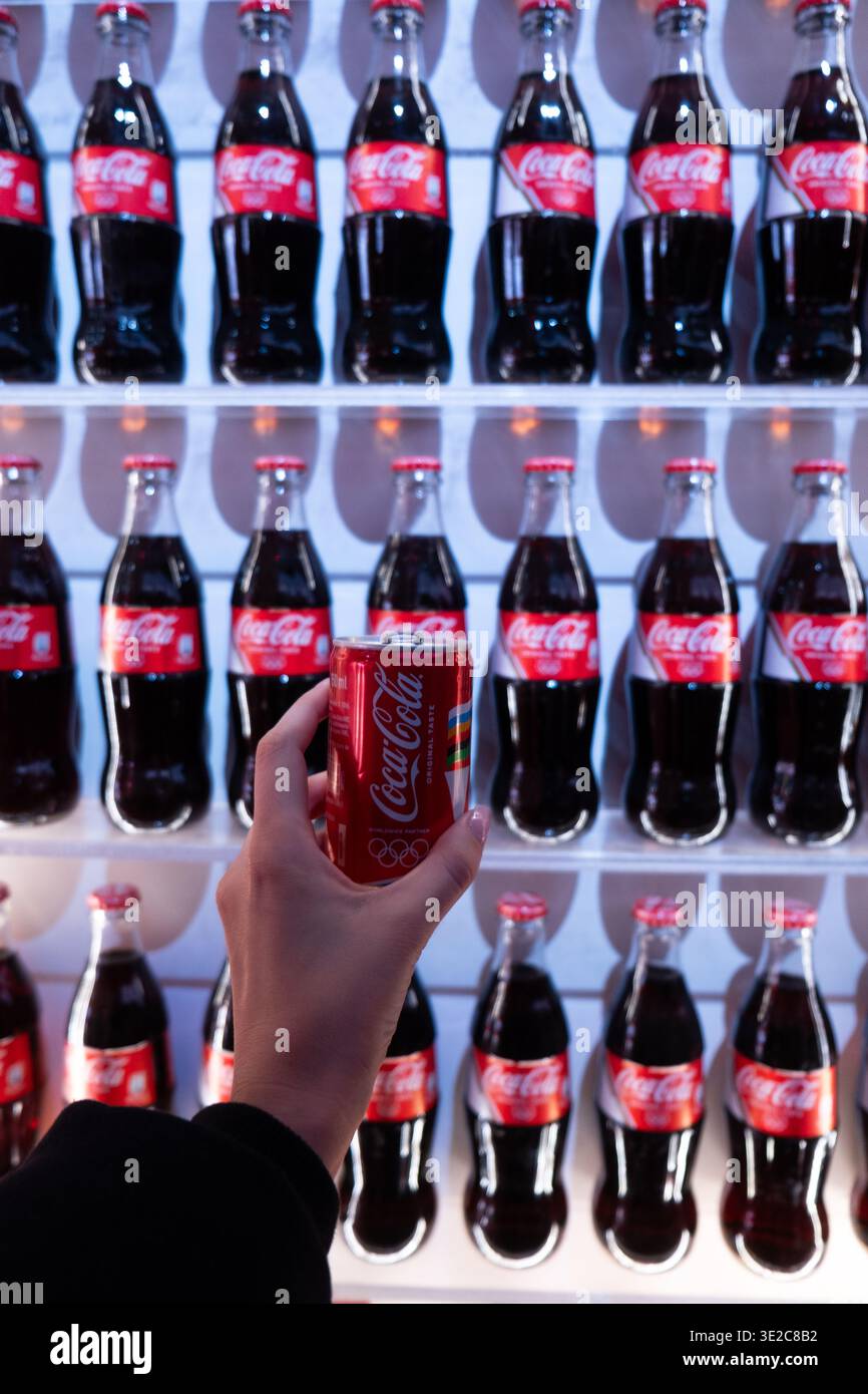 Coca-Cola-Dose in Handform vor dem Markendisplay mit Flaschenreihen, Werbeinstallation, Lifestyle-Konzept. Stockfoto
