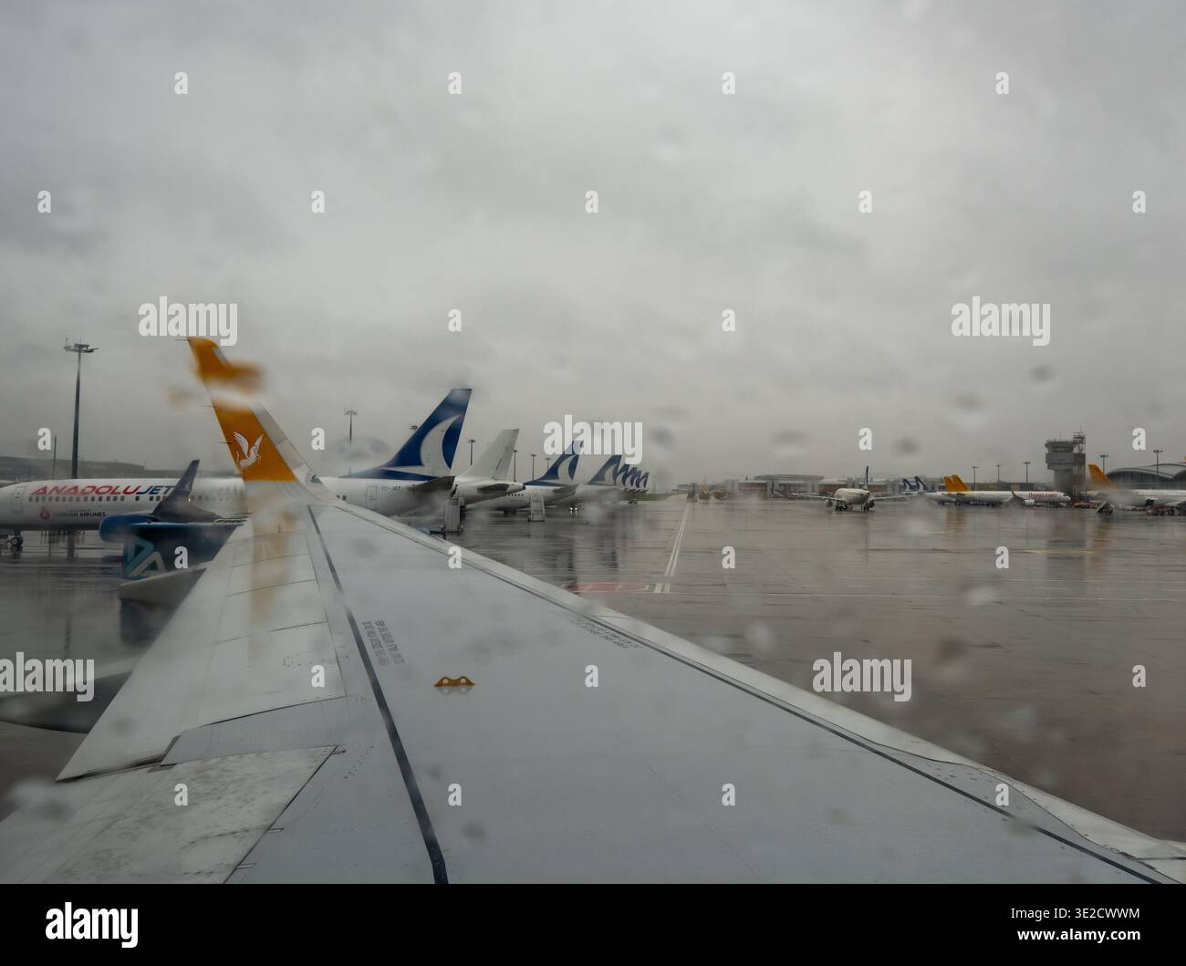 Blick aus einem Flugzeugfenster auf mehrere geparkte Passagierjets auf einem regnerischen Flughafenvorfeld mit nassem reflektierendem, asphaltierendem, grauem Himmel Stockfoto
