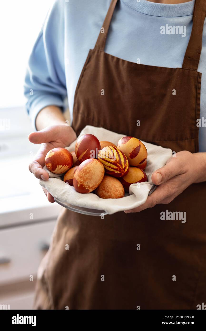 Person in brauner Schürze und hellblauer Oberseite hält eine Schüssel mit zwiebelgefärbten Ostereiern in einer hellen Wohnküche. Authentische Atmosphäre, Familien-Ostertrad Stockfoto