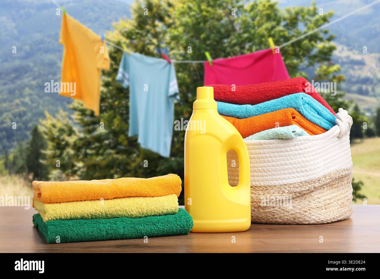Korb mit Wäsche und Flasche Waschmittel auf Holztisch in der Nähe von Kleidung, die draußen trocknet Stockfoto