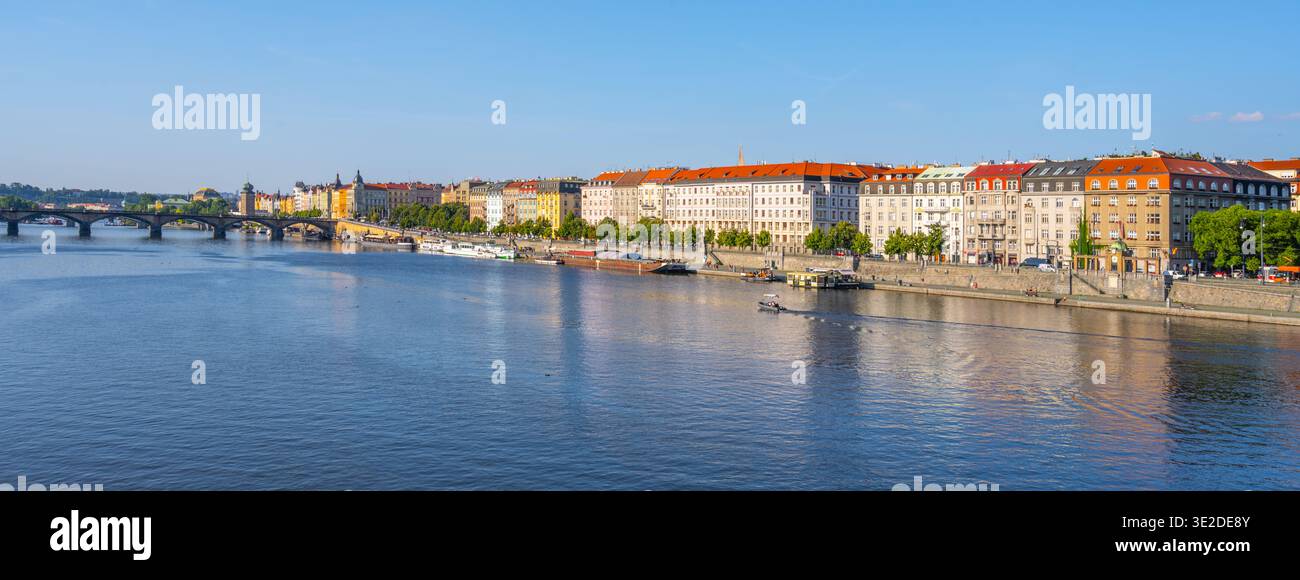 Die Menschen genießen ihre Zeit in der Nähe des Hafens Rasn in Prag. Die Sonne scheint hell, während sich die Boote langsam auf der Moldau bewegen. Gebäude säumen den Damm, gefüllt mit grünen Bäumen. Stockfoto