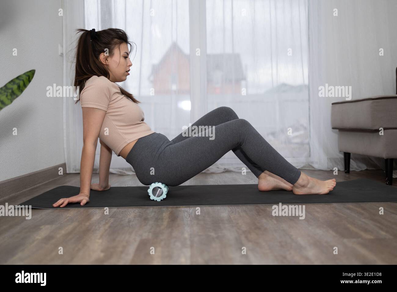 In einem Wohnzimmer sitzt eine junge Frau auf einer Yogamatte und benutzt eine Schaumstoffrolle auf ihrem Gesäß. Sie trägt Leggings und ein Hemd. Tagsüber strömt Licht aus einem Fenster. Stockfoto