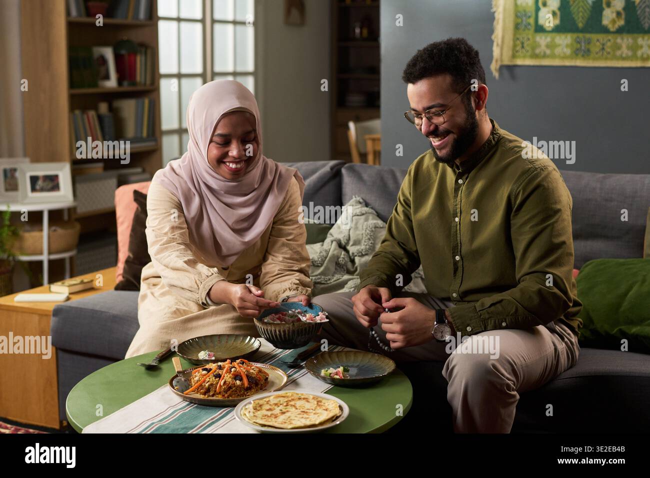 Junge Erwachsene muslimische Frau im Hijab und junger erwachsener muslimischer Mann, der auf dem Sofa sitzt und zusammen traditionelle Mahlzeiten teilt, sowohl lächelnd als auch mit Lebensmitteln interagierend Stockfoto
