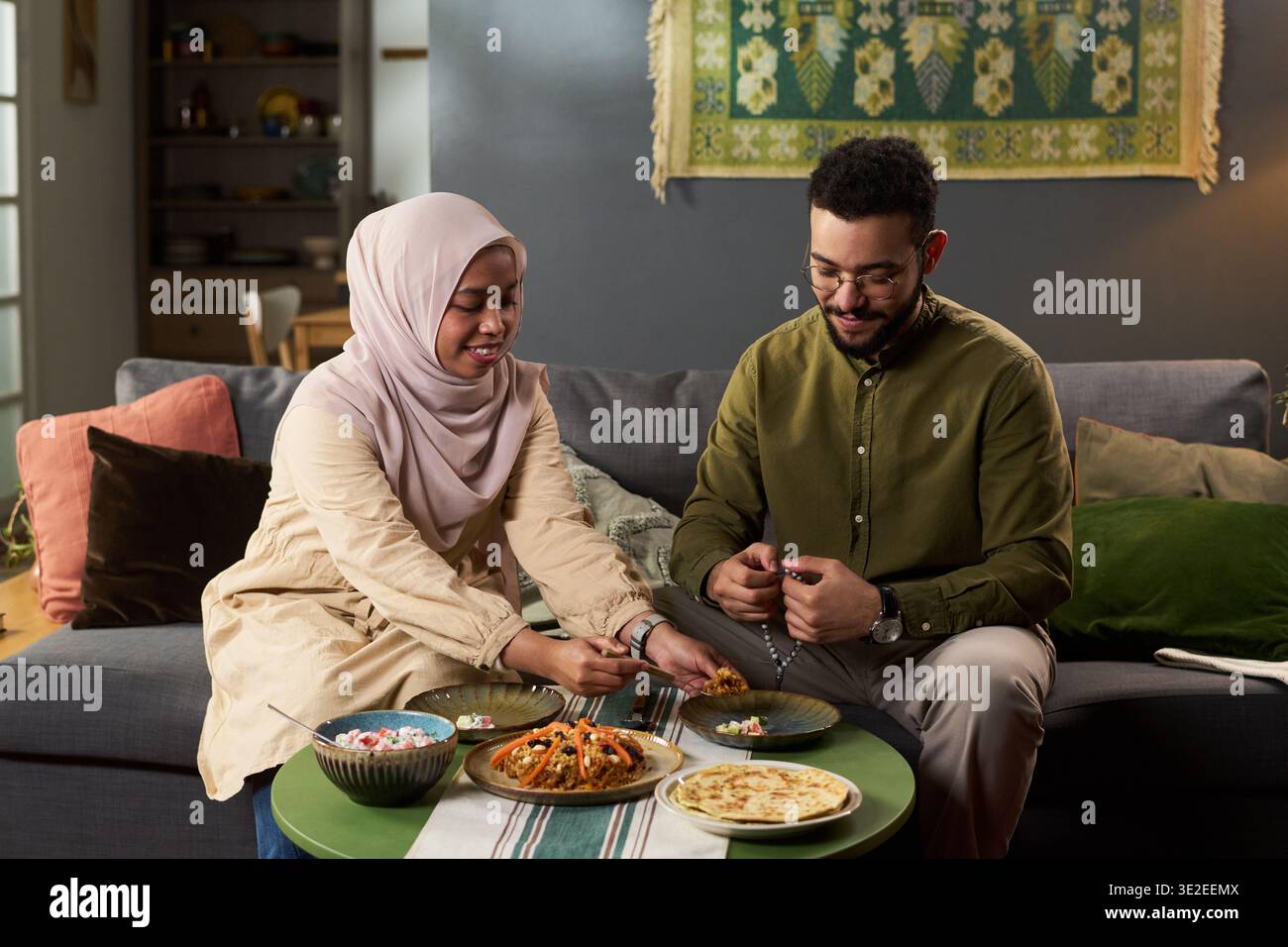 Junge Erwachsene muslimische Frau im Hijab und junger erwachsener Mann, der zusammen auf dem Sofa sitzt und zusammen Mahlzeiten teilt, lächelnd und genießt traditionelle Speisen im häuslichen Setting Stockfoto