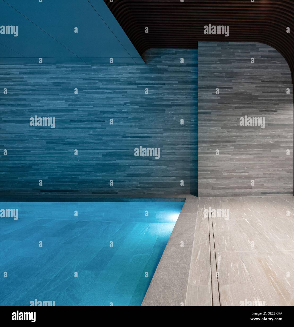 Hallenbad mit blauer Unterwasserbeleuchtung. Minimalistisches Spa-Interieur mit blauem Poolwasser, strukturierten Steinwänden und Holzdecke. Stockfoto