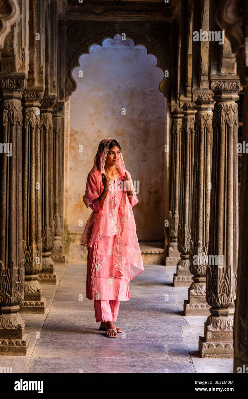 Eine Frau mit einem traditionellen rosa Salwar Kameez steht zwischen den kunstvoll geschnitzten Säulen einer historischen Kolonnade im Garh Palace in Bundi. Stockfoto
